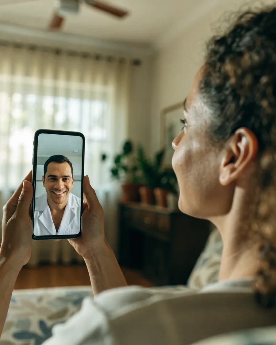 Paciente em casa segurando o celular numa videochamada com uma médica de jaleco branco